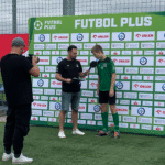 Na ściance z napisem „Futbol Plus” i logotypami partnerów wydarzenia dziennikarz rozmawia z zawodnikiem, naprzeciwko nich kamerzysta nagrywa materiał, po lewej na siatce ogrodzeniowej wisi baner Ministerstwa Sportu i Turystyki