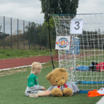 Na murawie boiska obok bramki z oznaczeniem nr 3 siedzi dziewczynka. Obok niej na trawie siedzi beżowy pluszowy miś prawie tak duży jak ona
