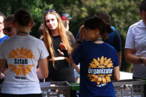 Na pierwszym planie dwie kobiety tyłem do aparatu. Od lewej kobieta w białej koszulce z napisem „Sztab”, z prawej w granatowej z żółtym logo i napisem „Organizator”