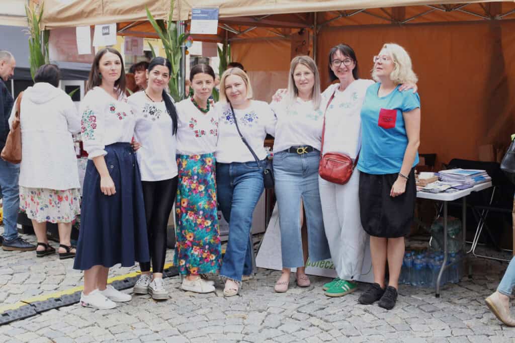 Siedem kobiet pozuje do fotografii na tle stoiska wystawienniczego. Kobiety obejmują się ramionami, uśmiechając się. Sześć kobiet od lewej ma na sobie białe bluzki z wyhaftowanymi kwiatami. Siódma z nich ma niebieską bluzkę