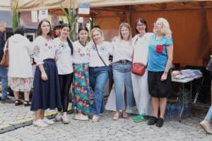 Siedem kobiet pozuje do fotografii na tle stoiska wystawienniczego. Kobiety obejmują się ramionami, uśmiechając się. Sześć kobiet od lewej ma na sobie białe bluzki z wyhaftowanymi kwiatami. Siódma z nich ma niebieską bluzkę