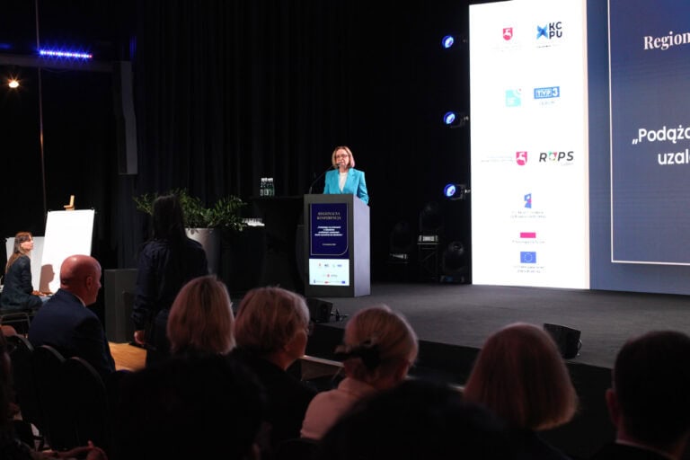 Wnętrze sali konferencyjnej, widok na scenę. Półmrok. Po lewej, przy mównicy kobieta (Małgorzata Romanko) podczas przemówienia. Z prawej fragment ekranu multimedialnego