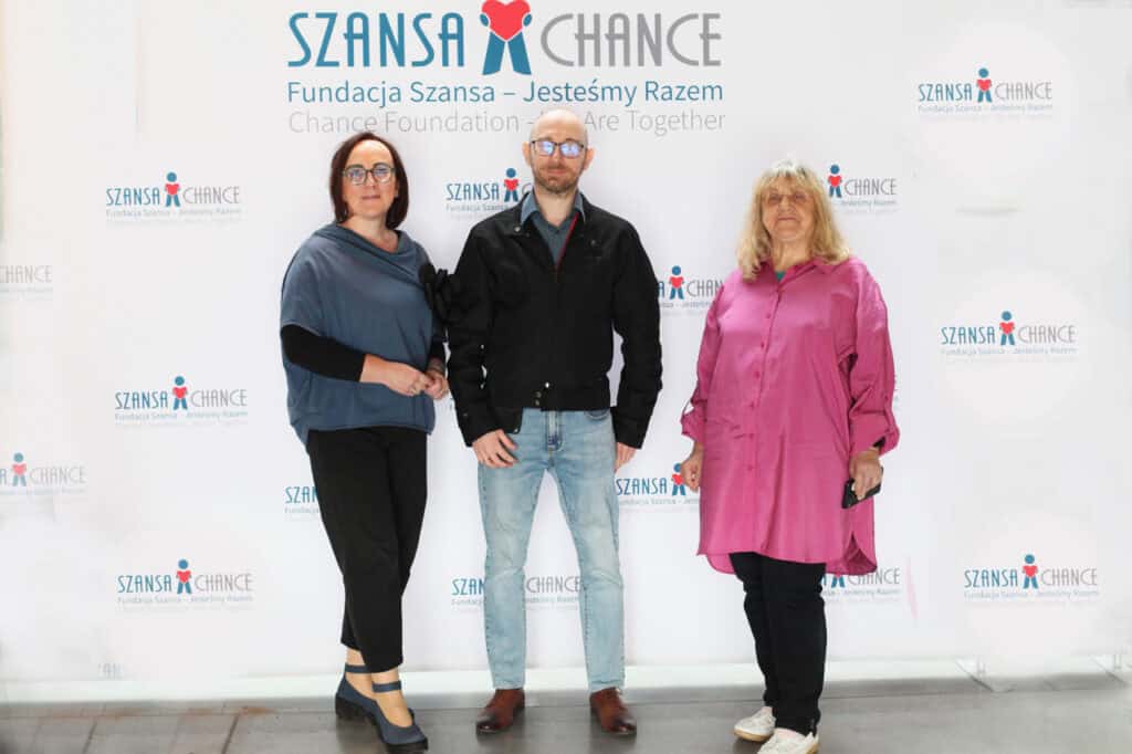 Na ściance fotograficznej z napisem: „Fundacja Szansa – Jesteśmy Razem” pozują do zdjęcia w planie pełnym trzy osoby. Od lewej: kobieta w okularach (Anna Bieganowska-Skóra - Prezes LFOON-SW), mężczyzna w okularach (Łukasz Lewicki - Wiceprezes LFOON-SW), kobieta w różowej koszuli (Alicja Jankiewicz - Prezes Honorowa LFOON-SW). Wszystkie osoby patrzą wprost do obiektywu
