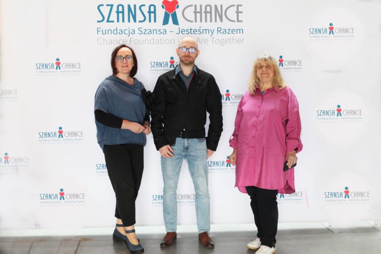 Na ściance fotograficznej z napisem: „Fundacja Szansa – Jesteśmy Razem” pozują do zdjęcia w planie pełnym trzy osoby. Od lewej: kobieta w okularach (Anna Bieganowska-Skóra - Prezes LFOON-SW), mężczyzna w okularach (Łukasz Lewicki - Wiceprezes LFOON-SW), kobieta w różowej koszuli (Alicja Jankiewicz - Prezes Honorowa LFOON-SW). Wszystkie osoby patrzą wprost do obiektywu