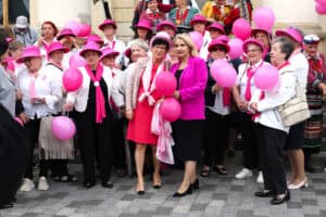 Plac Króla Władysława Łokietka 1 przed budynkiem Nowego Ratusza. Grupa kobiet w różnym wieku, ubranych głównie w różowe i białe stroje, w różowych kapeluszach i z różowymi balonami, pozuje razem na brukowanym placu. Dwie kobiety stoją z przodu, trzymając różowe balony. Tłum jest liczny, a na drugim planie widać część budynku