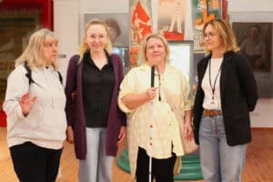 Pamiątkowa fotografia pozowana. Cztery kobiety pozują na tle słupa ogłoszeniowego oklejonego kolorowymi plakatami. W tle wnętrze sali wystawienniczej