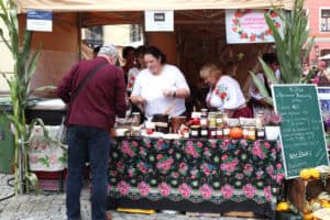 Stoisko Koła Gospodyń Wiejskich w Tarnawce Pierwszej. Na stoisku wyroby w słoikach z etykietami, gliniane gary. Obok zielona tablica z wypisanymi produktami, np.: flaczki, zupa dyniowa, pierogi z kapustą