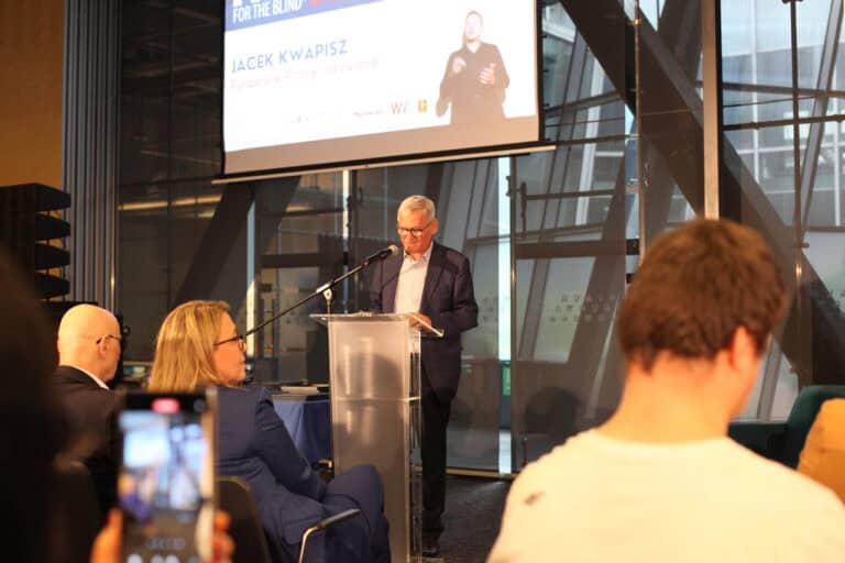 Mężczyzna w garniturze i okularach przemawia na podium do siedzącej publiczności w sali konferencyjnej. Za nim znajduje się ekran projekcyjny, na którym wyświetla się napis: „Jacek Kwapisz. Fundacja w Polsce i na świecie”