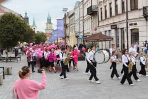Deptak w Lublinie. Parada maszeruje wzdłuż brukowanej ulicy miejskiej, z przodu której idzie młodzieżowa orkiestra dęta. Uczestnicy orkiestry są ubrani w czarno-białe stroje i niosą instrumenty. Za nimi podąża duża grupa ludzi, z których wielu jest ubranych w różowe ubrania i trzyma różowe balony. Po lewej stronie, na pierwszym planie, widać kobietę w różowym swetrze, która robi zdjęcie paradzie telefonem. W tle widoczne są zabytkowe kamienice i wieże