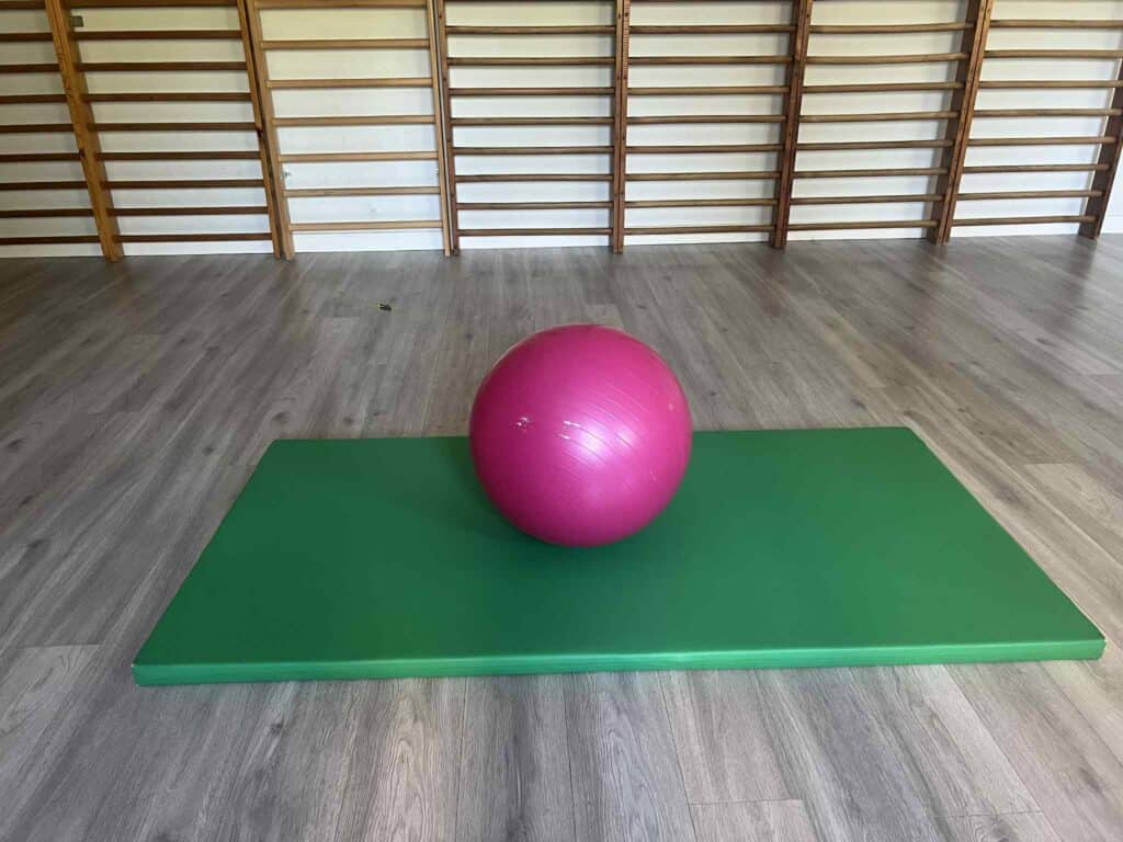 Na podłodze sali gimnastycznej leży zielony, cienki materac. Na materacu spoczywa ciemnoróżowa piłka rehabilitacyjna. W tle, przy ścianie zamontowane są drewniane drabinki gimnastyczne