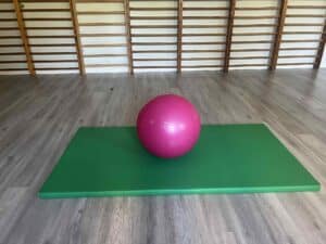Na podłodze sali gimnastycznej leży zielony, cienki materac. Na materacu spoczywa ciemnoróżowa piłka rehabilitacyjna. W tle, przy ścianie zamontowane są drewniane drabinki gimnastyczne