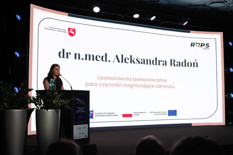 Kobieta przemawiająca na scenie. W tle duży ekran multimedialny, na którym wyświetlany jest napis: „dr n. med. Aleksandra Radoń. Uzależnienia Behawioralne jako czynniki zagrażające zdrowiu”