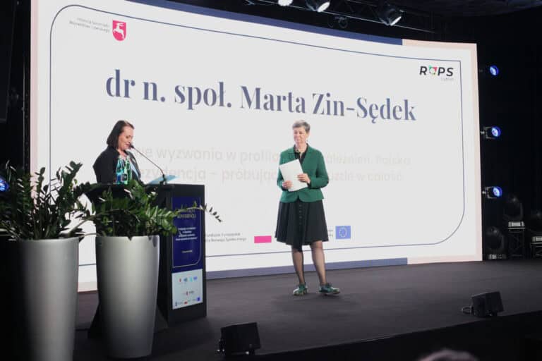 Dwie kobiety na scenie, jedna za mównicą, druga stojąca obok; w tle duży ekran multimedialny, wyświetlający tytuł "dr n. społ. Marta Zin-Sędek"