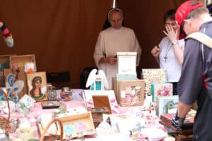 Przy stoisku z wyrobami rękodzieła artystycznego siostra zakonna i kobieta w białej bluzce z dużym krzyżem na szyi, poprawiająca dłonią okulary. Przed nimi, na stole wyroby artystyczne: ramki, ikony, anioły, kolorowe pudełka