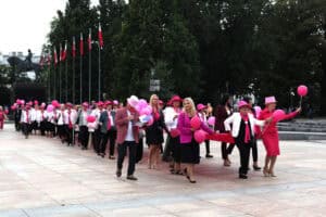 Plac Litewski w Lublinie. Grupa kilkudziesięciu osób maszeruje po placu miejskim wyłożonym płytami. Uczestnicy w większości są ubrani na różowo lub mają różowe elementy garderoby i balony. W tle widoczne są drzewa i szereg masztów z biało-czerwonymi flagami