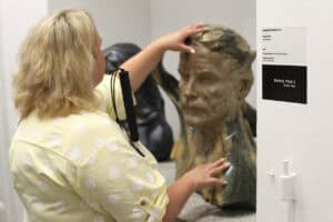 Od lewej: kobieta dotykająca dłońmi rzeźby w barwie brązu. Z prawej, na fragmencie ściany plakietka z napisem: „Dotknij i poczuj”