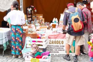 Stoisko Stowarzyszenia Osób Niepełnosprawnych Źródło, o czym świadczy tablica informacyjna stojąca przed stoiskiem. Przy stole wystawienniczym oglądający. Od lewej kobieta w białej bluzce i kolorowej spódnicy w kwiaty