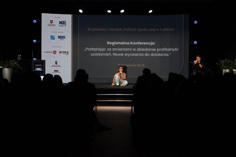 Sala konferencyjna z publicznością siedzącą w ciemności, na scenie siedzi mężczyzna z mikrofonem, a obok niego, z prawej siedzi tłumaczka języka migowego. W tle duży ekran z napisem "Regionalny Ośrodek Polityki Społecznej w Lublinie"