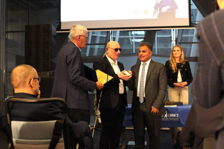 Ceremonia wręczania nagród na scenie. Dwóch mężczyzn w garniturach stoi pośrodku: jeden, w ciemnych okularach i trzymający białą laskę (Marek Kalbarczyk), wręcza mały przedmiot drugiemu mężczyźnie w szarym garniturze. Z lewej strony stoi mężczyzna wręczający dyplom. Z prawej strony w tle uśmiecha się kobieta. Na pierwszym planie, z lewej, widoczna jest osoba siedząca na wózku inwalidzkim