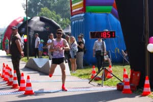 Linię mety przekracza mężczyzna w stroju sportowym, z czasem 18.51 widocznym na cyfrowej tablicy wyników. W tle kibice, namioty organizatorów. Od lewej linia biało pomarańczowych pachołków drogowych