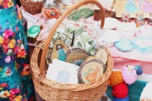 Wiklinowy koszyk z drewnianymi zawieszkami o różnych kształtach, na których namalowane są postacie i zwierzęta. W tle inne wyroby artystyczne