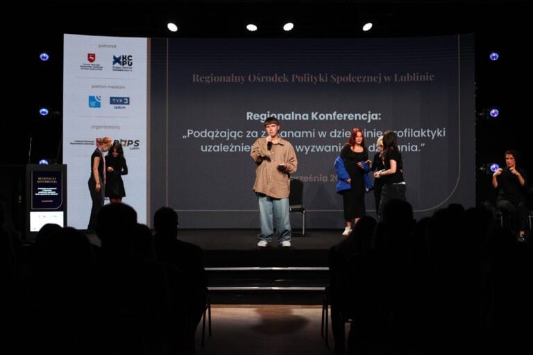 Sala konferencyjna z publicznością siedzącą w ciemności, na scenie stoi mężczyzna z mikrofonem, a obok niego, z prawej i lewej strony, grupki młodych kobiet