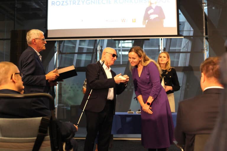 Na scenie odbywa się ceremonia. Mężczyzna w ciemnych okularach i z białą laską, ubrany w garnitur (Marek Kalbarczyk), uśmiecha się i wręcza mały przedmiot kobiecie w długiej, fioletowej sukience. Z lewej strony stoi mężczyzna trzymający dużą księgę. W tle, pod ekranem z napisem "ROZSTRZYGNIĘCIE KONKURSU", widoczna jest jeszcze jedna kobieta