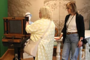 Od lewej aparat atelierowy wielkoformatowy, miechowy. Obok dwie kobiety, z których jedna dotyka dłońmi aparatu, druga obserwuje ją