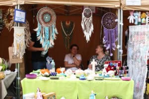 Stoisko Warsztatu Terapii Zajęciowej Niezwykli. Informuje o tym tabliczka z lewej. Trzy osoby za stołem z wyrobami artystycznymi. Do zadaszenia przymocowano kolorowe makramy i tzw. łapacze snów