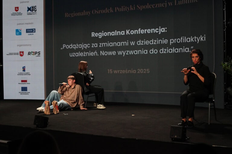 Widok na scenę pogrążoną w półmroku. Na scenie siedzą plecami do siebie mężczyzna i kobieta. Mężczyzna trzyma w ręku mikrofon. Z prawej tłumaczka języka migowego