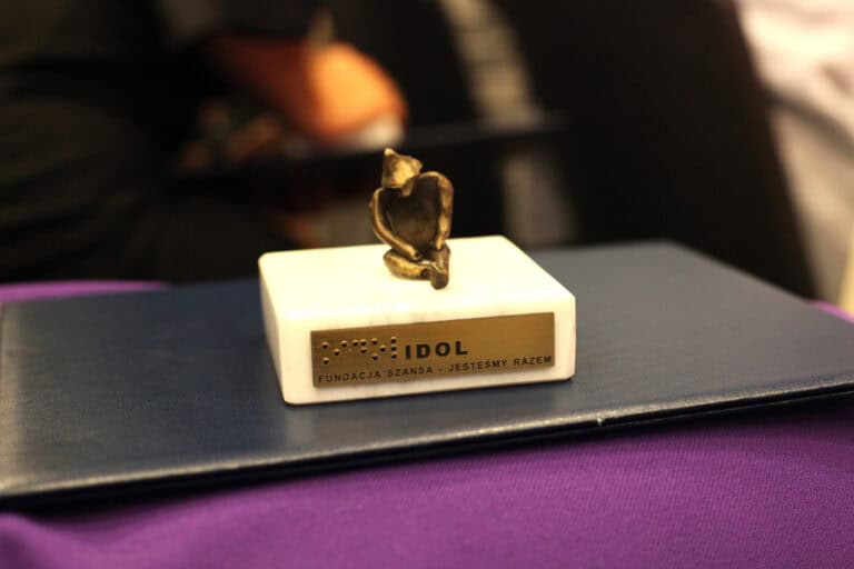 Zbliżenie na nagrodę IDOL umieszczoną na ciemnoniebieskiej teczce i fioletowym materiale. Nagroda to mała, metalowa figurka siedzącej osoby na prostokątnej, białej marmurowej podstawie. Na podstawie przymocowana jest złota tabliczka z napisem: czarnodrukiem "IDOL FUNDACJA SZANSA - JESTEŚMY RAZEM" oraz napisem w alfabecie Braille’a