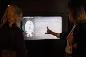 Półmrok. W centralnej części kadru ekran dotykowy z wyświetloną informacją o Aleksandrze Leszku Zamoyskim. Po obu jego stronach dwie kobiety, z których ta z prawej wskazuje coś palcem na ekranie