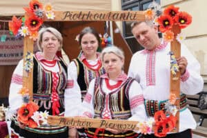 Cztery osoby pozują do zdjęcia w ramce do fotografii z napisem: Piotrkowianki, Piotrków Pierwszy. Trzy kobiety i mężczyzna ubrani są w kolorowe stroje ludowe