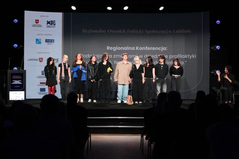 Sala konferencyjna z publicznością siedzącą w ciemności, na scenie pozuje do fotografii grupa młodych ludzi. Z prawej tłumaczka języka migowego