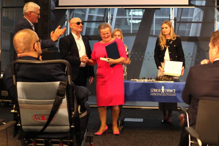 Na scenie odbywa się ceremonia wręczania nagród. Kobieta w różowej sukience i szpilkach uśmiecha się, trzymając w jednej ręce granatową teczkę, a drugą dłoń ściskając z mężczyzną w garniturze i ciemnych okularach (Markiem Kalbarczykiem). Mężczyzna po lewej klaszcze. W tle, za stołem z niebieskim obrusem, stoi inna kobieta trzymająca dyplom. Na pierwszym planie, z lewej strony, widoczna jest osoba siedząca na wózku inwalidzkim