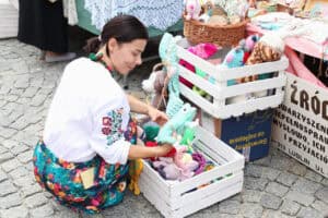 Kobieta w białej bluzce z kwiecistymi haftami na rękawach i kolorowej spódnicy pochyla się nad drewnianą skrzynią wypełnioną kolorowymi maskotkami w postaci zwierząt