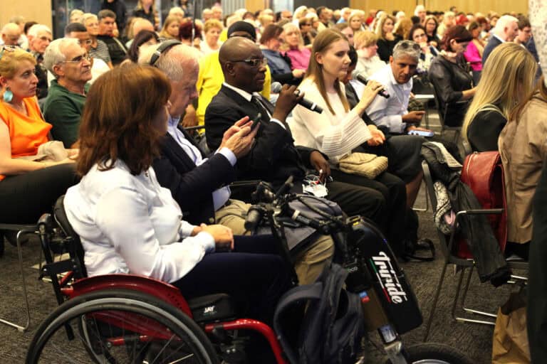 Ujęcie z bliska dużej publiczności siedzącej w rzędach podczas konferencji. Na pierwszym planie po lewej siedzi kobieta na wózku inwalidzkim, obok której siedzi mężczyzna w słuchawkach. Dalej, pośrodku, siedzi mężczyzna w garniturze i okularach, trzymający mikrofon. Kobieta obok niego również trzyma mikrofon