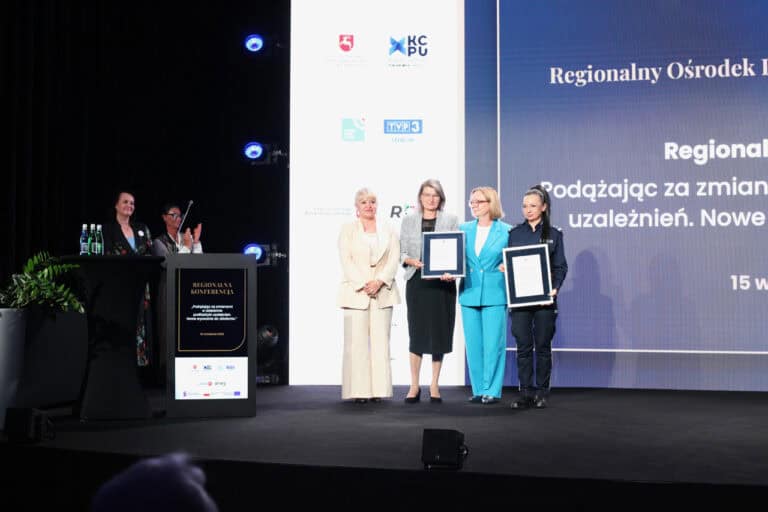 Grupa kobiet na scenie, w tym jedna w mundurze policyjnym. Kobiety pozują do pamiątkowej fotografii. Dwie z nich trzyma dokument oprawiony w ramkę