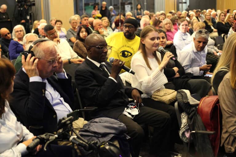 Ujęcie z bliska dużej publiczności siedzącej w rzędach podczas konferencji. Na pierwszym planie po lewej siedzi starszy mężczyzna w słuchawkach, a obok niego mężczyzna w garniturze trzymający mikrofon. Z prawej strony siedzi kobieta w białej bluzce, również z mikrofonem. W kolejnym rzędzie widoczny jest mężczyzna w żółtej koszulce z grafiką oka. Publiczność wypełnia całą salę