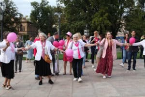 Plac Litewski w Lublinie. Grupa kobiet tańczy lub maszeruje, trzymając się za ręce. Uczestniczki są ubrane w stroje z różowymi akcentami, w tym różowe koszulki, kurtki, kapelusze i trzymają różowe balony. W tle zieleń drzew