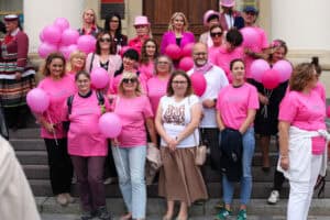 Plac Króla Władysława Łokietka. Grupa kobiet w różnym wieku, między nimi kilku mężczyzn. Kobiety ubrane są głównie w różowe koszulki. Pozują razem na schodach Ratusza. Część z nich trzyma różowe balony. W tle widać część budynku