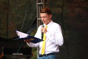 Mężczyzna w białej koszuli i żółtym krawacie. W prawej ręce trzyma otwarty program wydarzenia w twardej oprawie, w lewej ręce mikrofon. W tle nieostra zieleń drzew