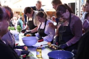 Rzut z boku na podłużny stół. Po obu jego stronach stoją kobiety w czarnych fartuchach kuchennych. Na plastikowych deskach kroją jabłka na cząstki