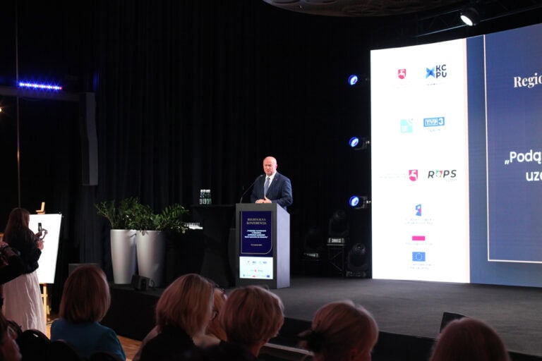 Wnętrze sali konferencyjnej pogrążone w półmroku. Na scenie, za mównicą mężczyzna (Jarosław Stawiarski) podczas przemówienia. Z prawej fragment ekranu multimedialnego