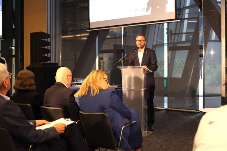 Mężczyzna w garniturze i okularach przemawia na podium do siedzącej publiczności w sali konferencyjnej. Za nim znajduje się ekran projekcyjny z niewyraźną prezentacją