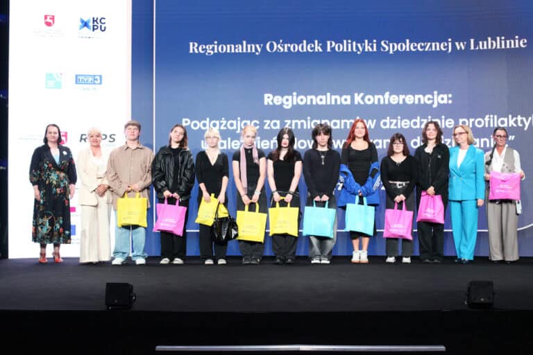 Grupa dwunastu osób, w tym młodzież i dorośli, stojąca na scenie i trzymająca kolorowe torby. W tle duży ekran wyświetlający nazwę "Regionalny Ośrodek Polityki Społecznej w Lublinie"