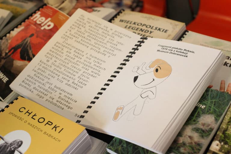 Zbliżenie na otwartą książkę leżącą na stosie innych książek. Po prawej stronie widoczny jest rysunek psa Reksia (postaci z kreskówki) z podpisem w czarnodruku: "Fragment plakatu Reksio, 1978 rok z kolekcji Muzeum Dobranocek". Po lewej stronie znajduje się tekst o Reksiu oraz numer strony. Strony książki zawierają zarówno czarnodruk, jak i tekst w wypukłym brajlu.