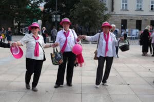 Plac Litewski w Lublinie. Trzy kobiety pozują do zdjęcia trzymając się za ręce. Kobiety mają na sobie białe bluzki i różowe elementy garderoby, takie jak apaszki, daszki, wstążki. W tle miejska architektura i zieleń drzew