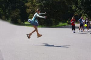 Kobieta na wrotkach, w sukience gimnastycznej, uchwycona w podskoku, w trakcie robienia piruetu. Na asfaltowym podłożu cień kobiety. W tle zieleń drzew. Z prawej strony kadru kilkuosobowa grupa oglądająca występ