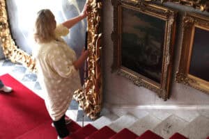 Widok z góry. U podnóża schodów stoi kobieta z białą laską. Dotyka opuszkami palców grubej, złotej, zdobionej ramy okazałego obrazu. Obok, na prawo mniejszy obraz w zdobionej ramie i fragment kolejnego. Na schodach widać brzeg czerwonego dywanu